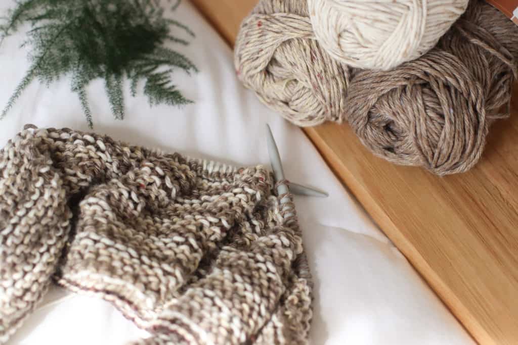 three skeins of neutral colored yarn on a wooden background, circular knitting needles with knitting. Tatamy Tweed worsted yarn
