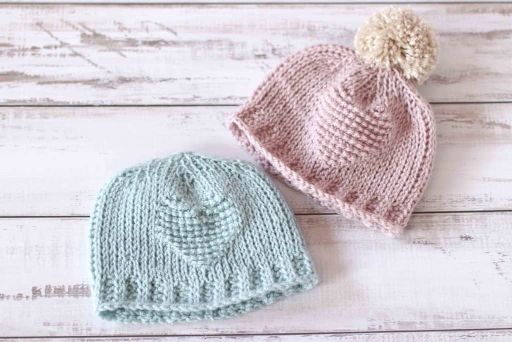 Two Tunisian crochet baby hats in pink and mint green, with heart embellishment.