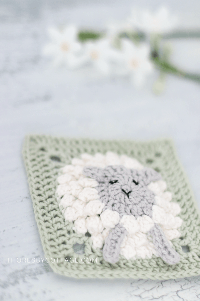Fluffy white lamb, green crocheted square with flowers in the background. 