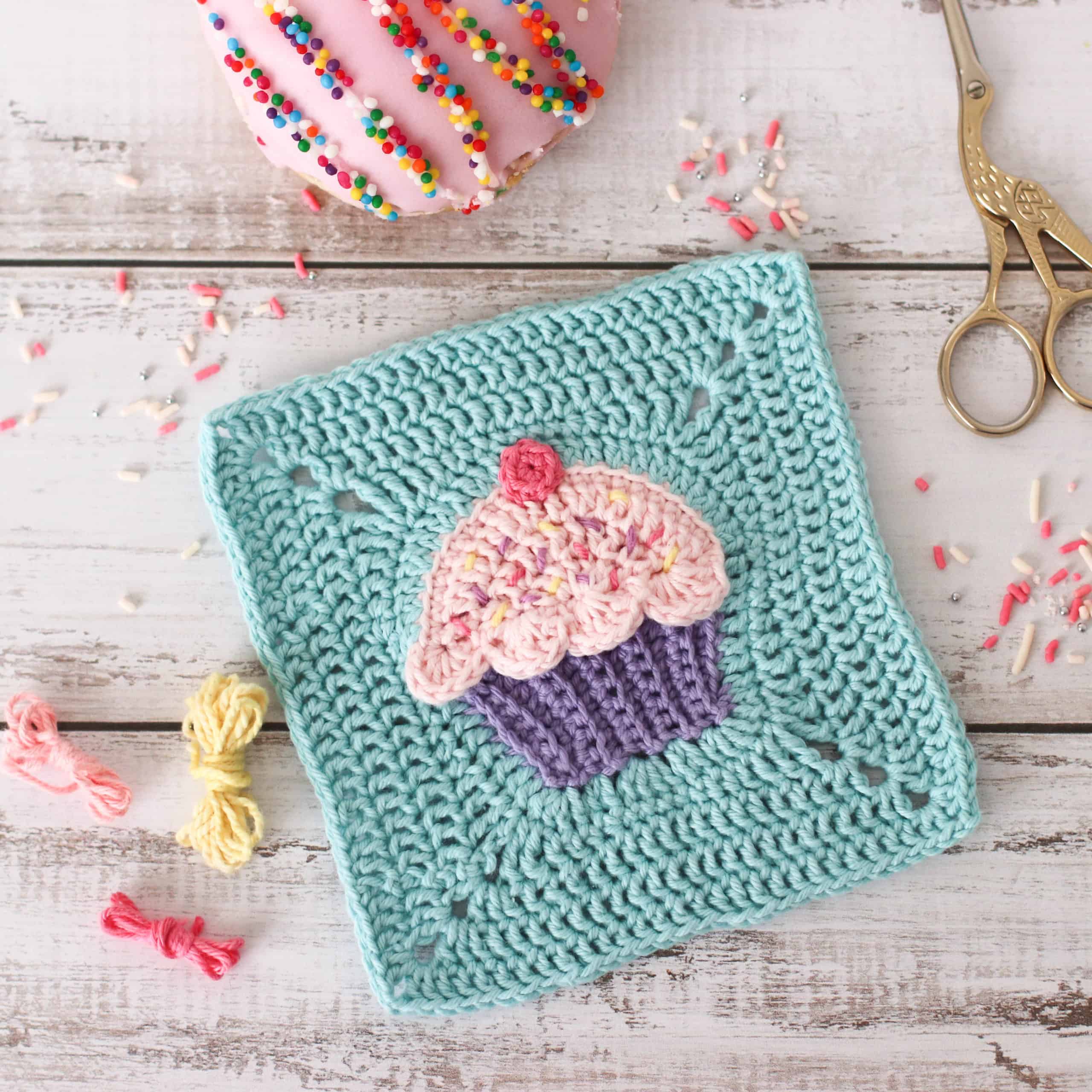 pink crochet cupcake square with a blue background, surrounded by sprinkles, yarn and donuts.