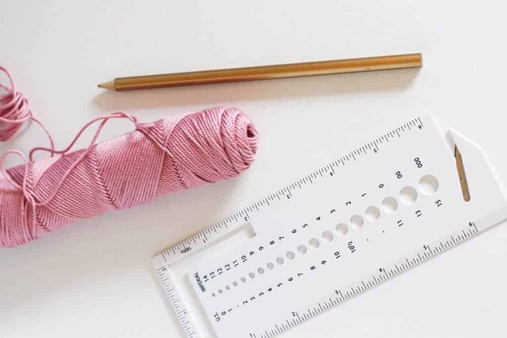 Pink cotton yarn next to a gold pencil and white gauge tool