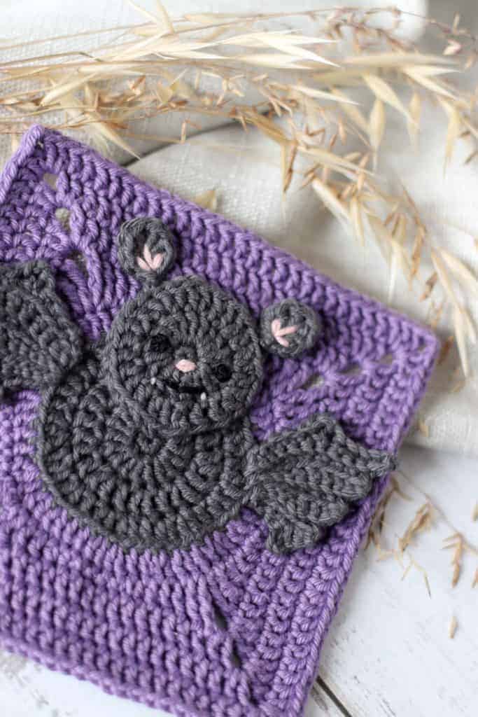 crochet bat pattern | grey crochet bat in a purple granny square, surrounded by linen and grass seeds